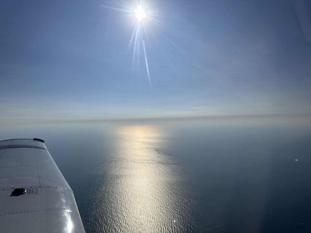 Airplane wing over ocean, sun shining bright.
